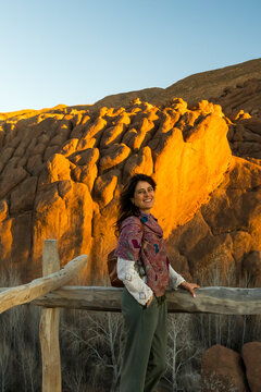 A smiling woman in a patterned shawl stands by a wooden fence with a rock formation behind her called Monkey fingers. Dades gorges,Monkey fingers,Marrakech,Morocco