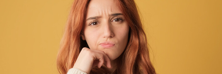 Fototapeta premium Close-up, pensive red-haired woman dressed in sweater thinks something, dreams on yellow background. Beautiful girl poses in studio with blank space on yellow wall for your promotion. Panoramic