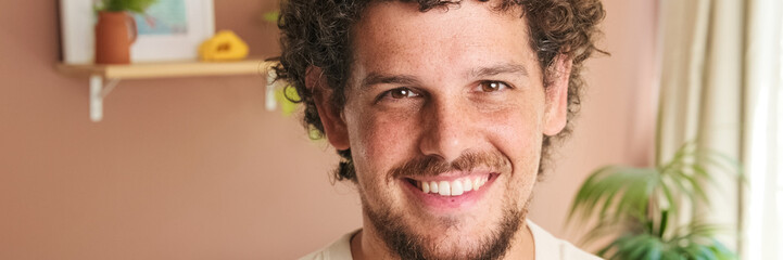 Obraz premium Close-up of young man with curly hair and beard looking at the camera with smile while standing in cozy living room. Panoramic