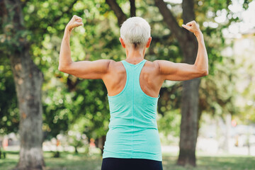 Senior woman flexing arms in park wearing teal fitness top outdoors in nature highlighting health...