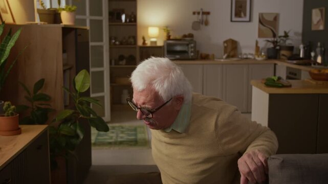 Medium shot of senior 80-year-old Caucasian man struggling to move sofa at home, then collapsing on floor and crying with intense back pain, while suffering from geriatric arthritis in old age
