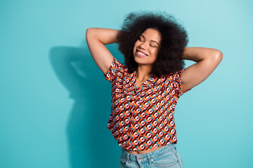 Young woman with curly hair smiles with arms behind head wearing a colorful patterned shirt against a blue background