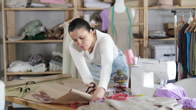 Smiling young woman seamstress cutting pieces of paper with scissors according to drawn template for pattern during dress design at workplace in modern sewing studio