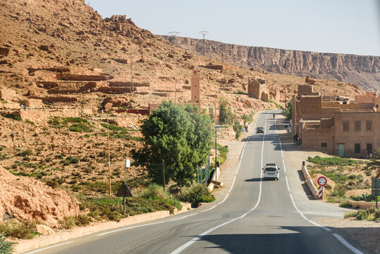 Paved desert road winding through the sandstone cliffs of Ziz Valley, Morocco, with small buildings scattered along the slope