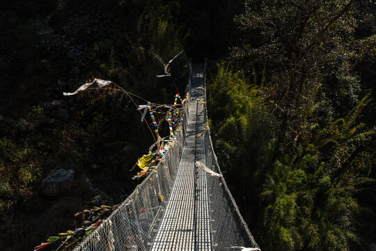Narrow suspension bridge with fluttering prayer flags crossing deep forest gorge in Langtang Valley, Nepal, seen from a moody elevated angle