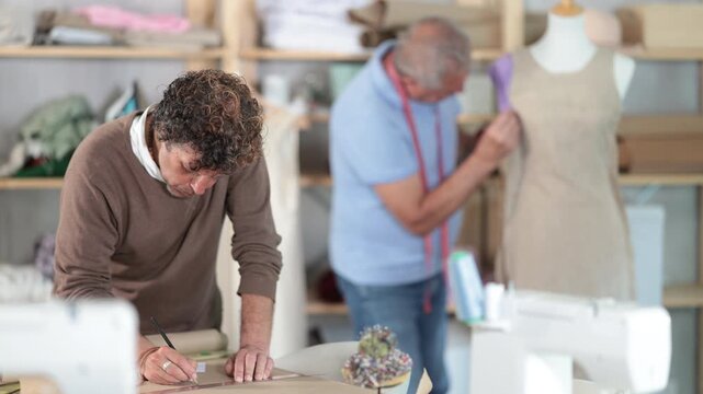 Male clothing designer drawing a sketch of clothes on paper using the client's individual measurements against the background of a tailor in a sewing workshop. High quality 4k footage