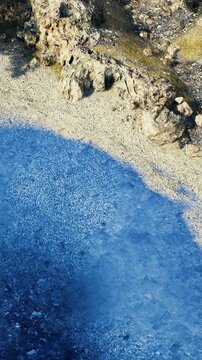 aerial pebble beach with azure shallow water and jagged rocks, textured shoreline gradient, gentle surf ripples and bright coastal light, tranquil natural