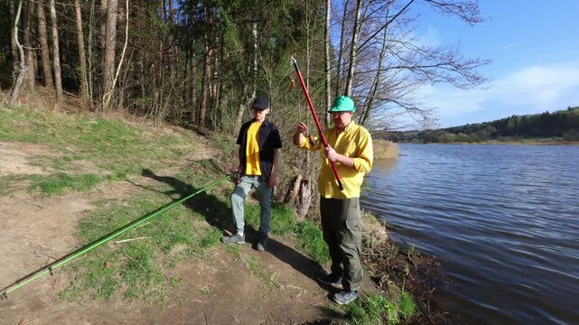 Two individuals stand by a lakeside, preparing fishing rods amidst a serene forest backdrop.