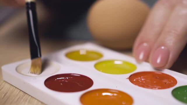Close-up of woman hands takes white paint for coloring easter eggs with painting brush. Holidays, tradition and people