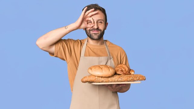 Smiling male baker in apron holding assorted breads and making a positive gesture on blue background