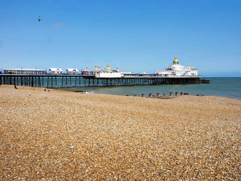Visitors walk on shingle beach near pier in Eastbourne with clear skies and calm waters during daytime in East Sussex, United Kingdom