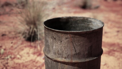 Fototapeta premium A rusty, weathered barrel is positioned on arid ground surrounded by scattered dry grasses. The dry setting suggests a remote location, possibly in a rural area.