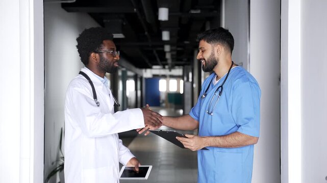 Two doctors standing in hospital corridor talking and shaking hands after discussion. Multiethnic male medical professionals smiling after successful discussion, trust, respect, positive cooperation