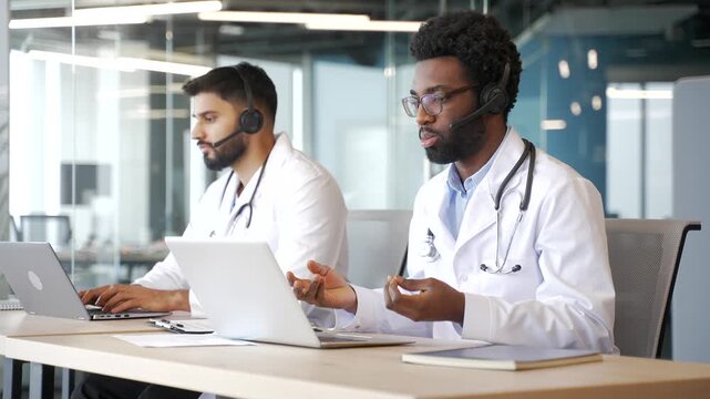 Two male doctors in white coats with headsets working on laptops in medical call center. Multiethnic healthcare professionals helping patients remotely, offering consultation and advice in office.