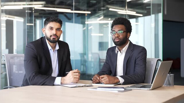 Portrait of two businessmen in suits sitting at office desk with serious confident expressions. Multiethnic colleagues looking focused and determined, leadership, professionalism and business strength