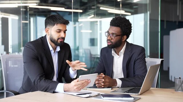 Two businessmen sitting at office desk using digital tablet during meeting. Multiethnic colleagues in formal suits discussing project data, planning successful business strategy in corporate workspace