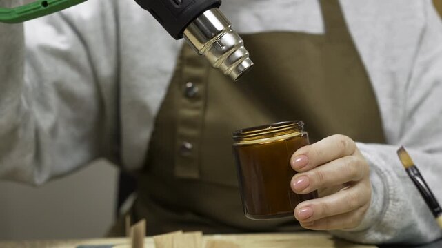 female hands melting wax in homemade candle with professional construction dryer indoors