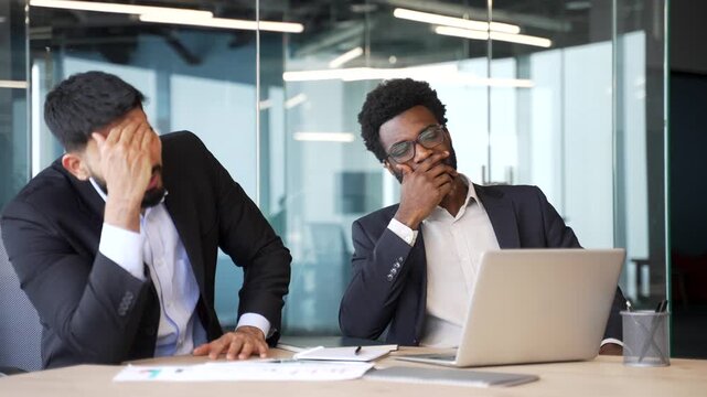 Two businessmen in suits sitting at office desk looking at laptop with disappointed expressions. Multiethnic colleagues showing frustration and sadness after failure or bad results in corporate office