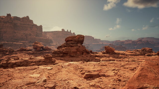 Sunlight bathes the rugged terrain in a desert landscape. Large rock formations rise majestically as soft clouds drift overhead, creating a serene yet dramatic atmosphere.