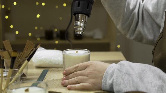 Close-up of female hands melting wax in homemade candle with professional construction dryer indoors