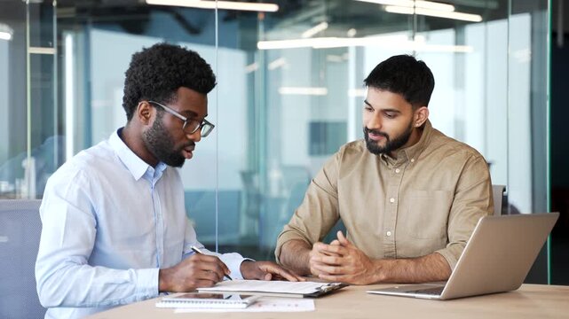 Sales manager sitting at office desk signing contract with client and shaking hands after successful deal. Multiethnic business partners confirming agreement, cooperation and trust in modern workspace