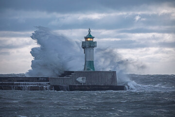 Fototapeta premium Leuchtturm Sassnitz bei Wintersturm – riesige Brecher schlagen an die vereiste Kaimauer, leuchtendes Signallicht in dramatischer Ostsee-Gischt auf Rügen