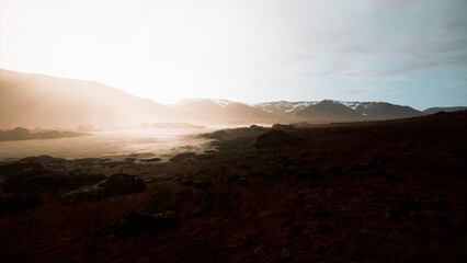 Golden light breaks over distant mountains, illuminating a rocky terrain. Mist lingers low, creating a serene atmosphere. Nature awakens as day begins in this tranquil setting.