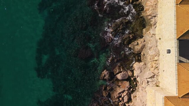 Epic top down aerial pedestal up reveal of orange roofs. 
Cinematic vertical flight panorama of historic medieval city walls, 
rocky beach, sea waves foam and blue ocean water in Croatia.
