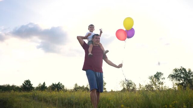 Father and son in field holding balloons. Balloons with a man and boy. A woman is walking outdoors with her shoulder bounding in the field. A dad and his son are in a field carrying lifestyle.