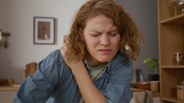 Medium close-up of mature Caucasian woman in denim shirt massaging sore shoulder and neck muscles after experiencing sharp pain or cramp, while doing routine household chores in kitchen