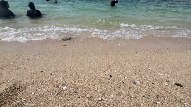 "Gentle ocean waves rolling onto a white sandy beach with people swimming in the clear turquoise water in the background."