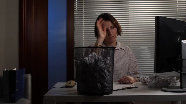 Woman sitting at an office desk, looking frustrated and throwing a crumpled piece of paper into a full waste bin, symbolizing creative block, stress, and failed attempts by an employee working late