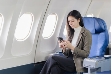 Fototapeta premium An Asian woman sits in a seat on an airplane and talks on her smartphone. A business travel concept.