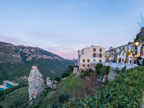 View of a rock formation below a building with red shutters, blending with the rugged mountains and a serene lake at dusk, El Castell de Guadalest, Valencian Community, Spain.