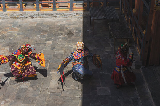 Jakar Dzong, Bhutan - 24 September 2025: View of masked dancers in vibrant costumes performing on the stone courtyard, shadowed by the dzong's walls, creating an intense contrast of light and dark.