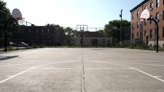 An empty outdoor basketball court with white markings on concrete, surrounded by chain-link fencing and red brick apartment buildings on a bright sunny day.