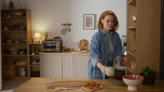 Medium shot of middle-aged Caucasian woman making lunch in kitchen, taking dishware out of cupboard, reaching for top shelf, getting injured by falling basket, rubbing forehead and grimacing in pain