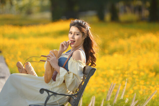 Young woman in a romantic cottagecore Renaissance type dress sitting in a chair in a field of yellow flowers eating a baguette, Thailand