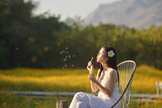 Side view of a young woman sitting in a chair blowing bubbles with a bubble wand at sunset, Thailand