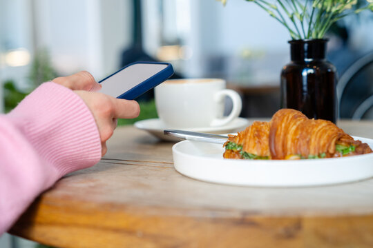 Close-up side view of a woman sitting at a table using her mobile phone with a cup of coffee and croissant with a fried egg and greens