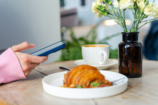 Close-up side view of a woman sitting at table in a coffee shop using her mobile phone with a cup of coffee and Savoury Croissant filled with greens and a Poached Egg on the table