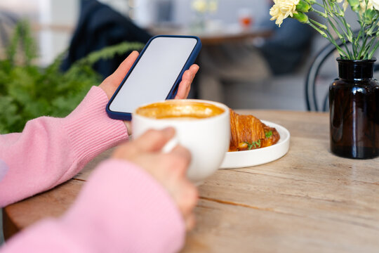Close-up of a woman sitting at a table drinking a cup of coffee while holding a mobile phone with a blank screen