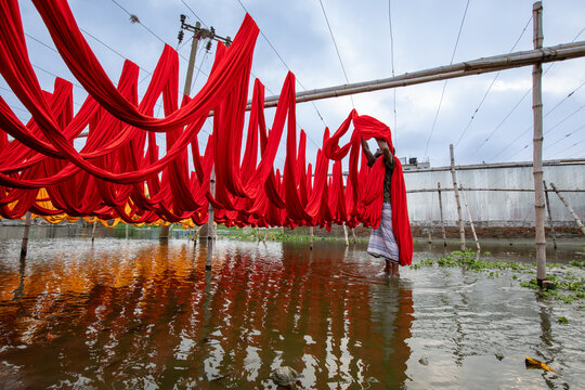 Narayanganj, Bangladesh - 05 October 2021: View of vibrant red and yellow fabrics suspended above the water, mirroring the sky, as a figure tends to the cloth, creating a striking contrast.