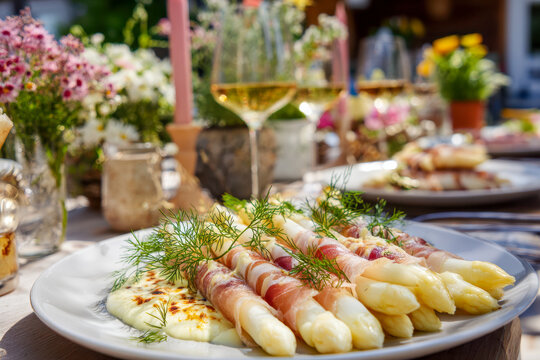 Elegant outdoor dining setup featuring creamy baked white asparagus wrapped in cured ham and garnished with fresh herbs alongside glasses of white wine and floral de