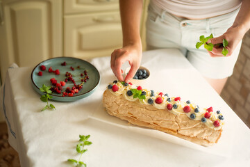 process of making homemade meringue roll cake decorated with fresh berries and mint. female hands adding raspberries on top, cozy kitchen atmosphere, baking at home concept.