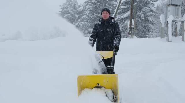 A middle-aged man is removing snow with a snow blower in a strong blizzard.  4K Resolution slow motion footage.