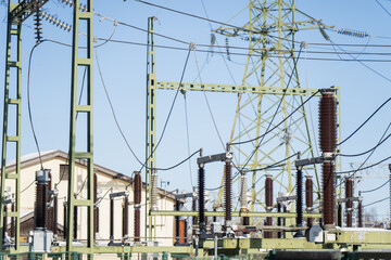 Utility power grid switchyard showing porcelain insulators, busbars and wiring on green metal frames, with lattice transmission pylon behind, industrial infrastructure