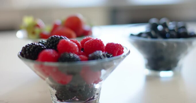Camera shifting focus centering glass bowl of raspberries  and  blackberries, background bowls blurr