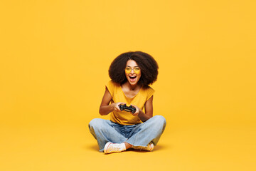 Full body young woman of African American ethnicity wear t-shirt casual clothes sitting hold in hand play pc game with joystick console isolated on plain yellow orange background. Lifestyle concept. © ViDi Studio