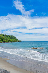 Obraz premium Tropical coastline with lush green forest, rocky shore, and gentle ocean waves under a bright blue sky in Indonesia.